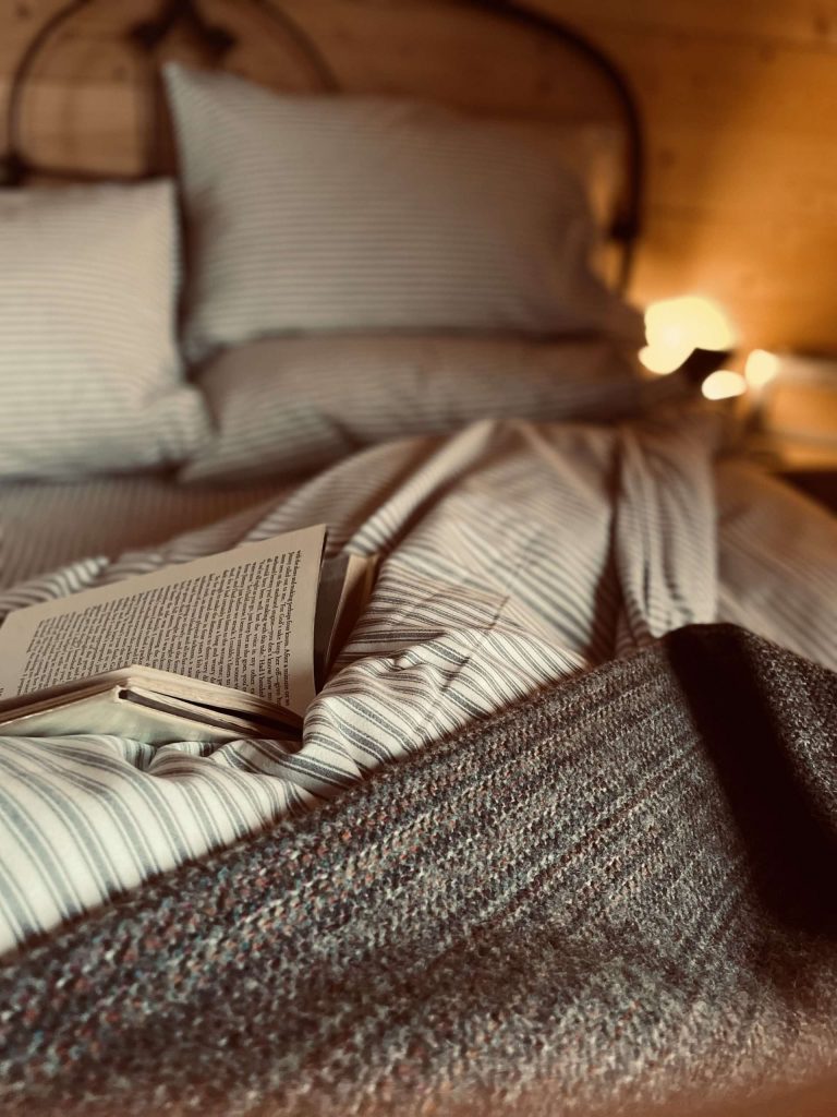 A stylistic shot of the bed, the bedding is purposefully messy with an open book and moody lighting, evocative of a night spent reading. 